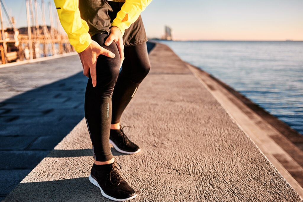 A runner holding his knee in pain near the waterfront, representing knee osteoarthritis and sports-related knee injuries treated by Dr. Burke Orthopedics in Houston.